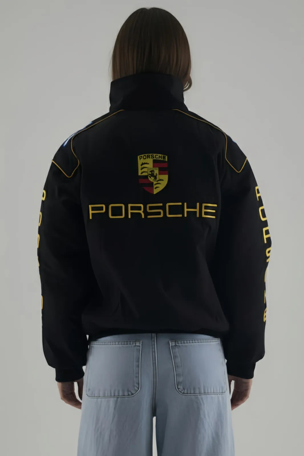 Female Model wearing a black Porsche jacket with logo and text on a plain background giving Back pose
